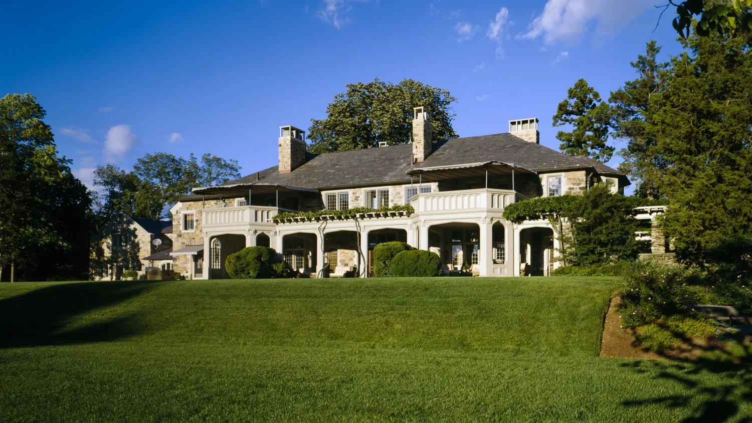 Large house with stone exterior, multiple chimneys, and a wrap-around porch, surrounded by green lawn and trees, under a blue sky.