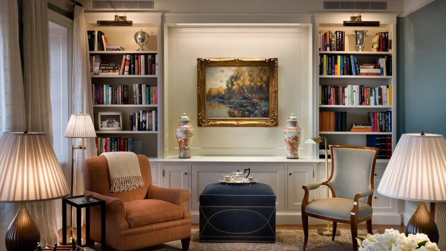 Living room with built-in white bookshelves, a framed landscape painting, table lamps, an upholstered armchair, a chaise lounge chair, a dark blue ottoman, decorative vases, and a tray with a tea set.