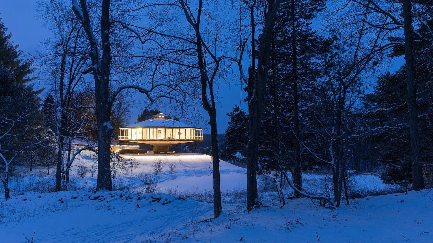 A modern house with large windows illuminated from within, sitting on a snowy landscape surrounded by leafless trees at dusk.