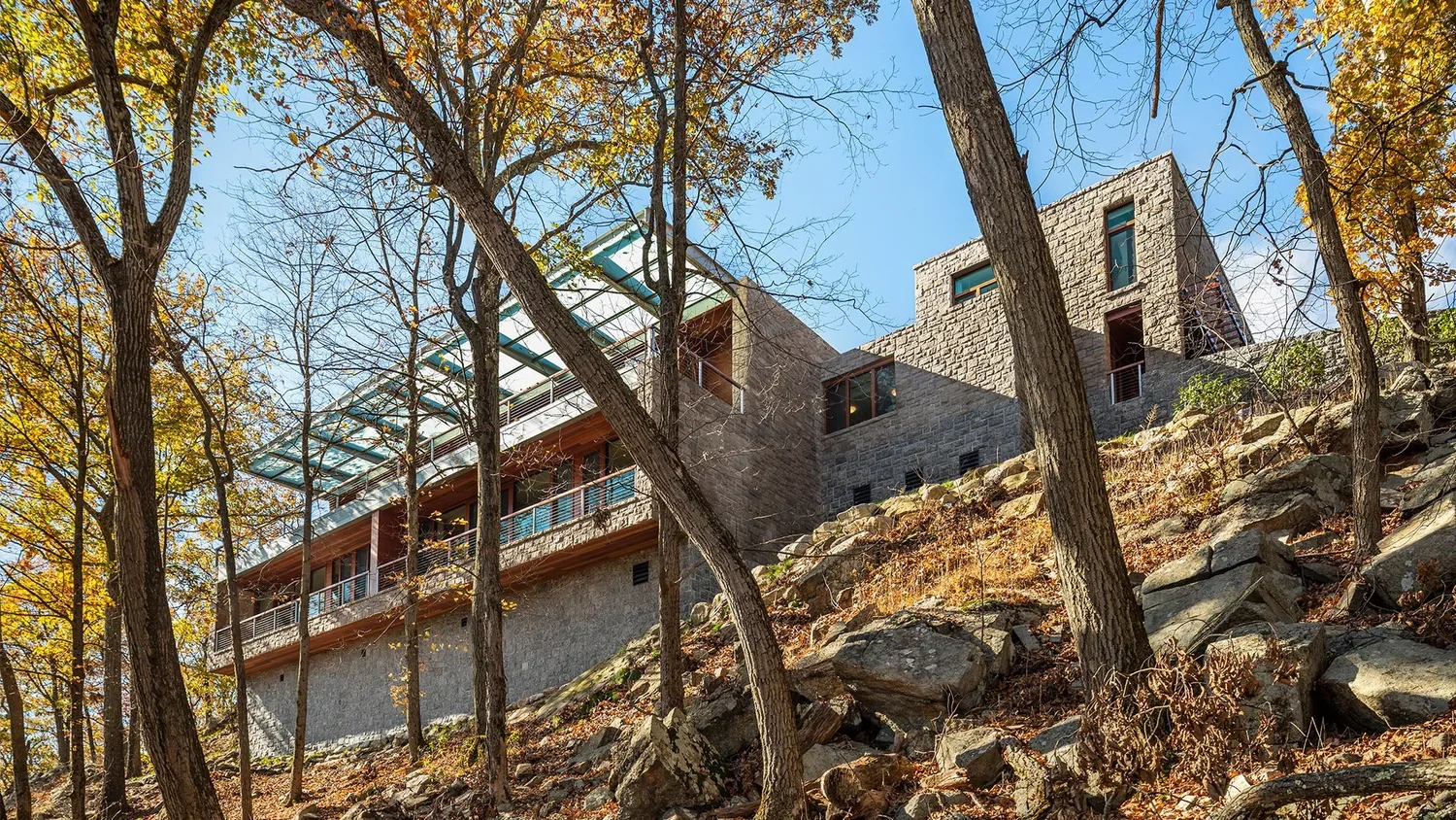 Modern house built on a rocky hillside surrounded by trees with changing autumn leaves, under a clear blue sky.