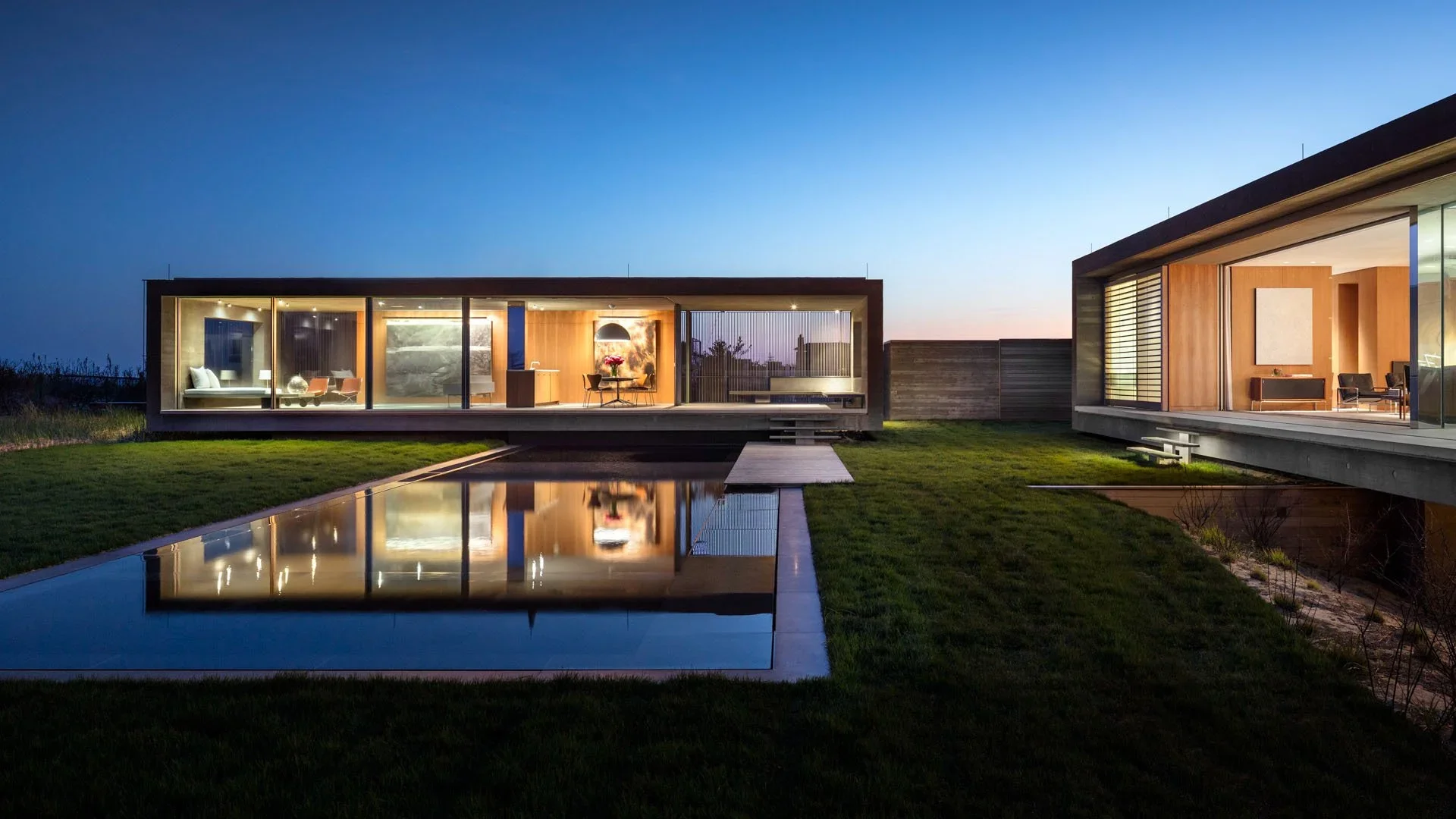 Modern house with large glass windows, overlooking a reflecting pool and landscaped lawn during dusk.