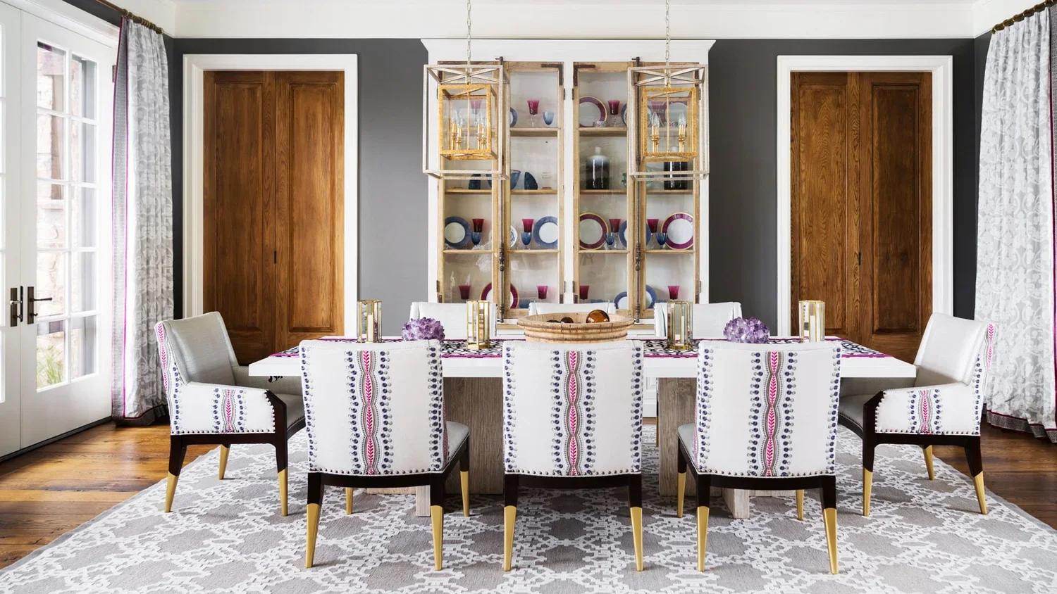 A modern dining room with a white table, ten patterned chairs, wooden doors, a display cabinet with colorful dishes, and hanging pendant lights.