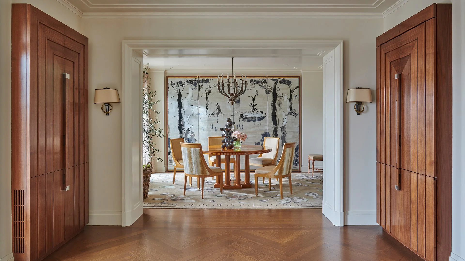 View of a dining room through an entryway with wooden cabinets, wall sconces, and a chandelier, featuring a round wooden table with a vase of pink flowers and sculptures, a large abstract black and white artwork on the wall, and a patterned rug.