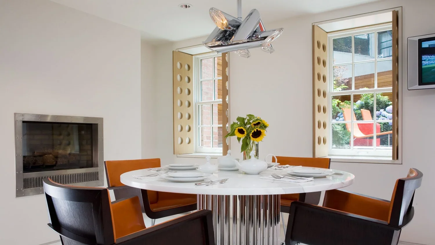 Modern dining room with a round white marble table, four chairs with black and brown leather, a vase with sunflowers, windows with wooden shutters, and an abstract silver sculpture ceiling light.