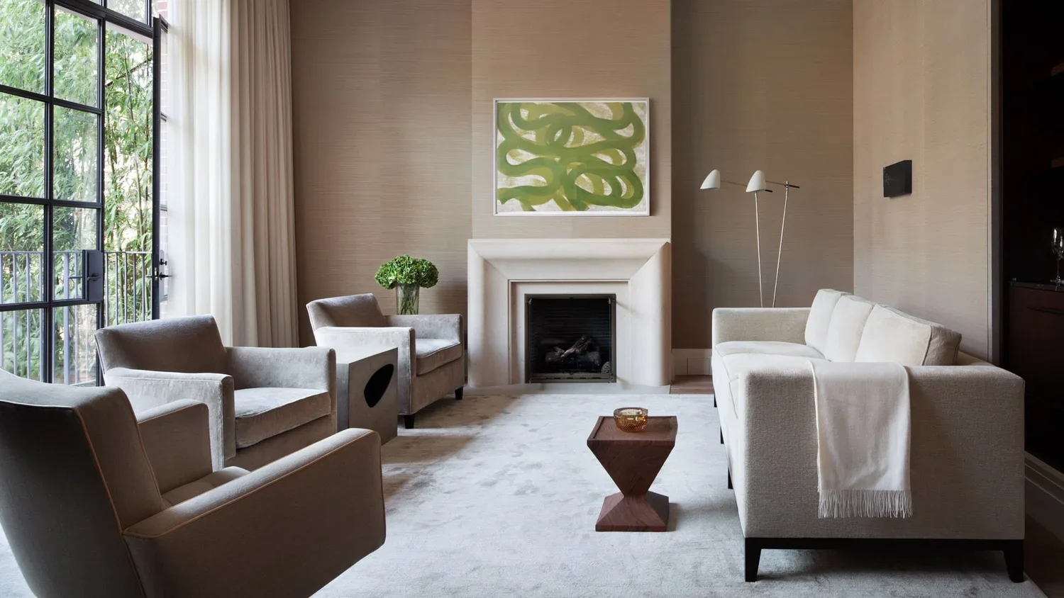 A modern living room with beige and white furniture, a fireplace, a green abstract painting above the fireplace, and large windows with white curtains.
