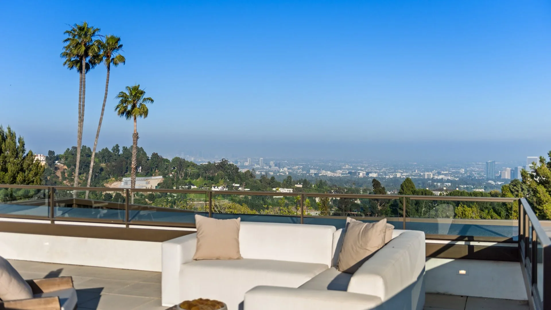 A rooftop patio with white outdoor furniture and beige pillows, overlooking a city skyline with tall buildings, palm trees, and green hills under a clear blue sky.