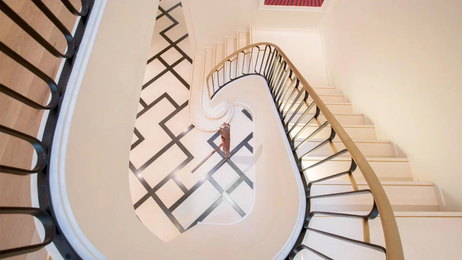 Top view of an indoor staircase with black railing, white steps, and black-and-white geometric patterned floor at the bottom.