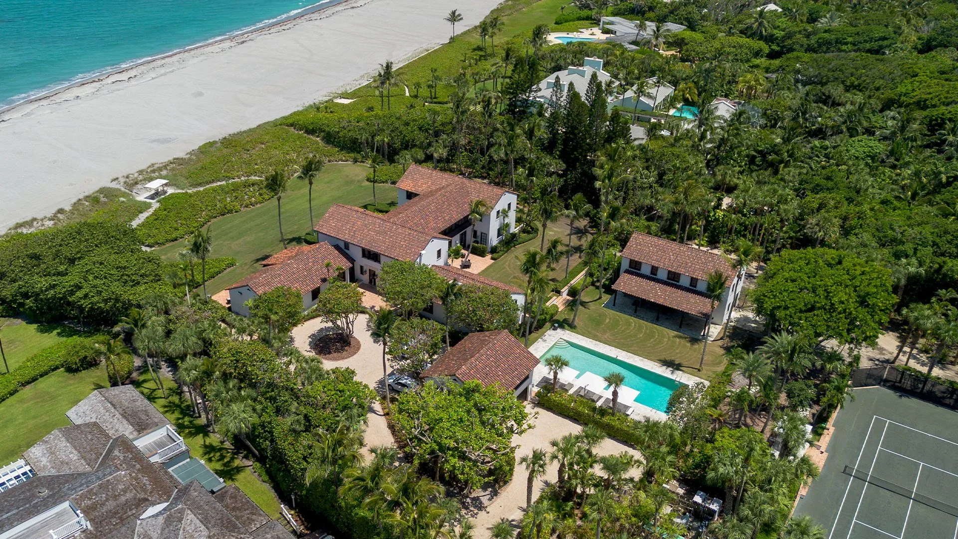 Aerial view of a luxurious beachfront property with multiple white buildings, swimming pool, lush green landscaping, tennis court, and sandy beach shoreline.