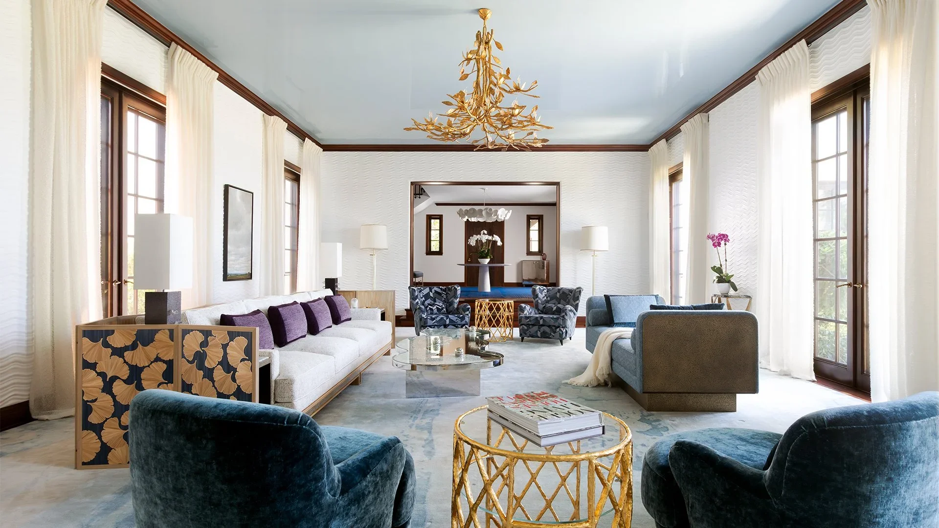 Living room with large windows, white curtains, modern furniture, a chandelier, and decorative accents.