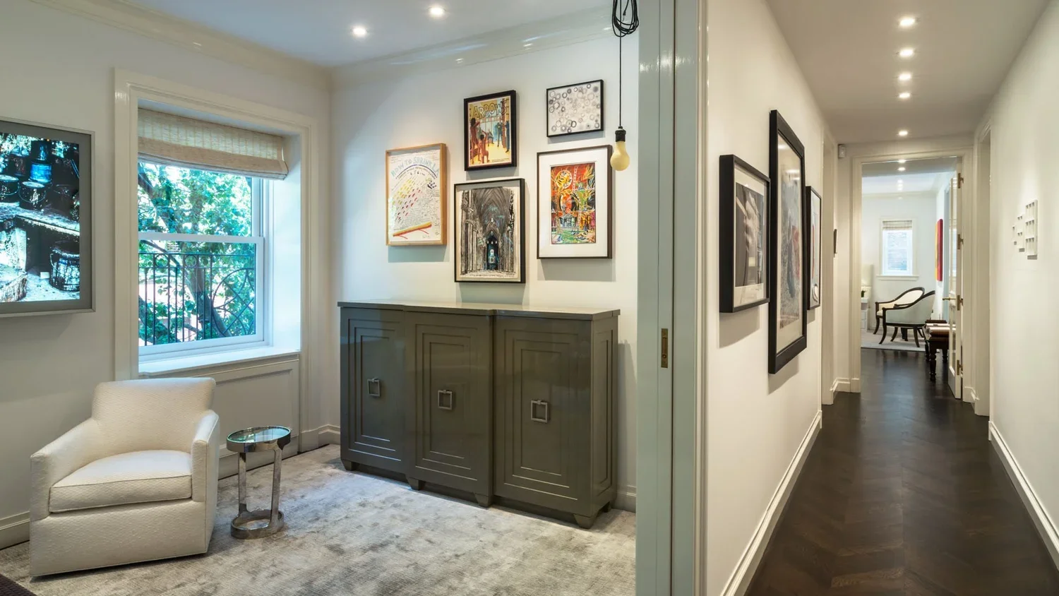 A hallway leading to a living room with dark wooden flooring, white walls, and multiple framed pictures on the walls. The living room features a window with a beige shade, a small white armchair, a silver side table, and a dark green cabinet. Natural