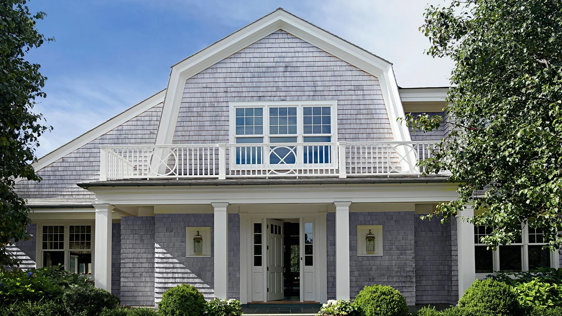 Front view of a house with gray shingle siding, white trim, a balcony with a white railing, a gabled roof, and landscaped bushes and trees.