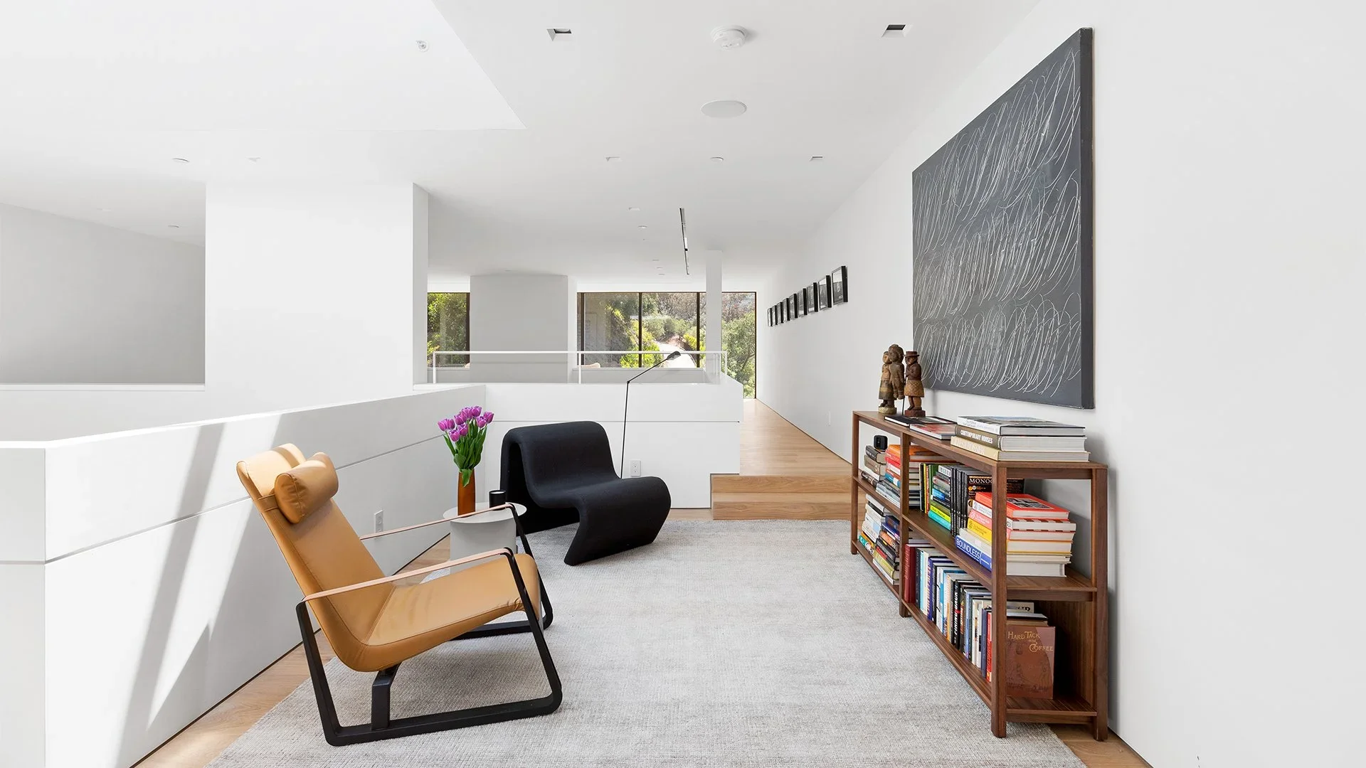 Modern living room with white walls, a beige lounge chair, a black armchair, a wooden bookshelf filled with books, a vase of pink tulips, artwork on the walls, and large windows with a view of greenery outside.