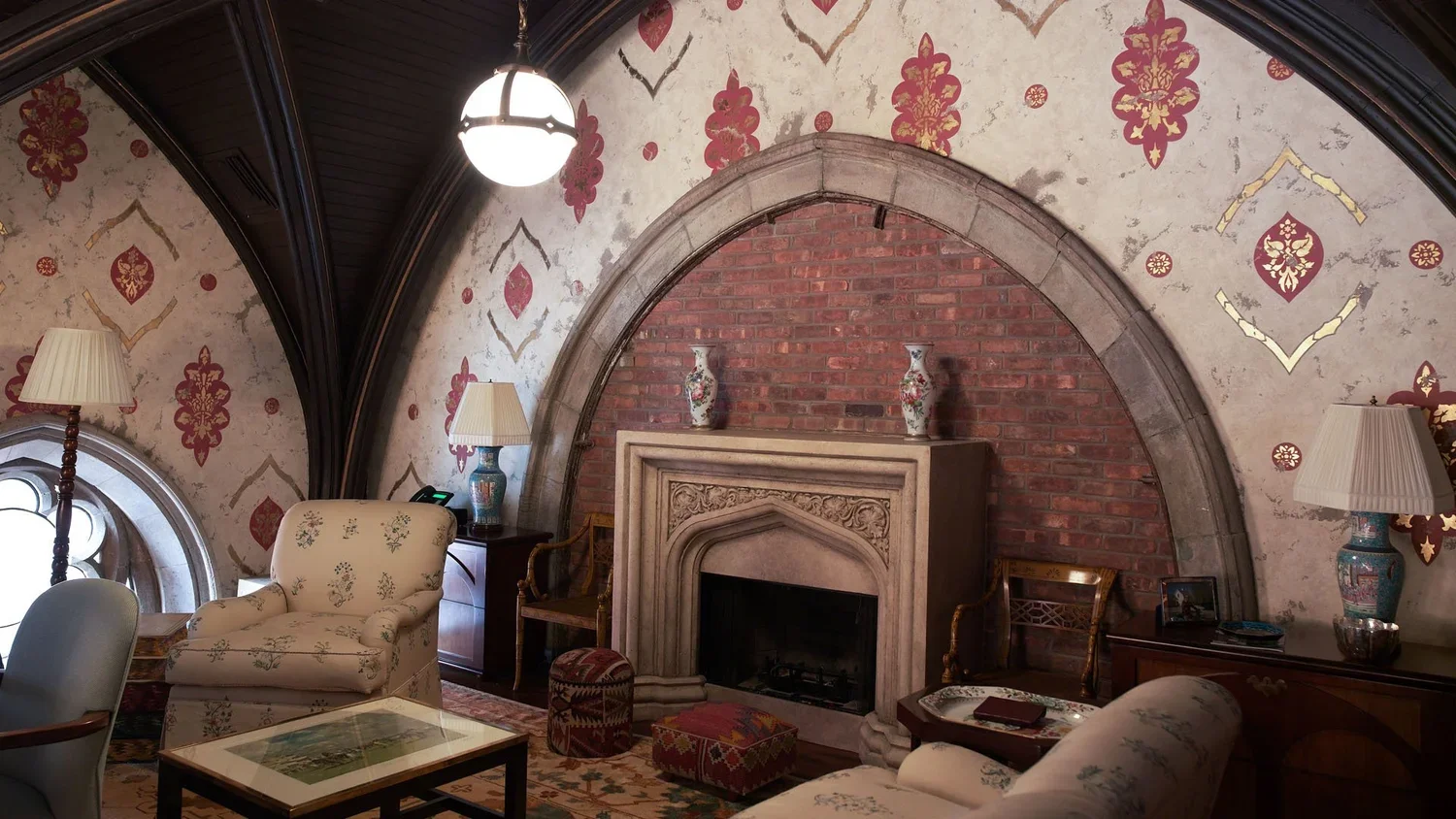 A cozy living room featuring a brick fireplace with a detailed stone mantel, surrounded by patterned wallpaper with red and gold accents, and decorated with vases on the mantle. The room includes vintage-style furniture such as armchairs, lamps, and 