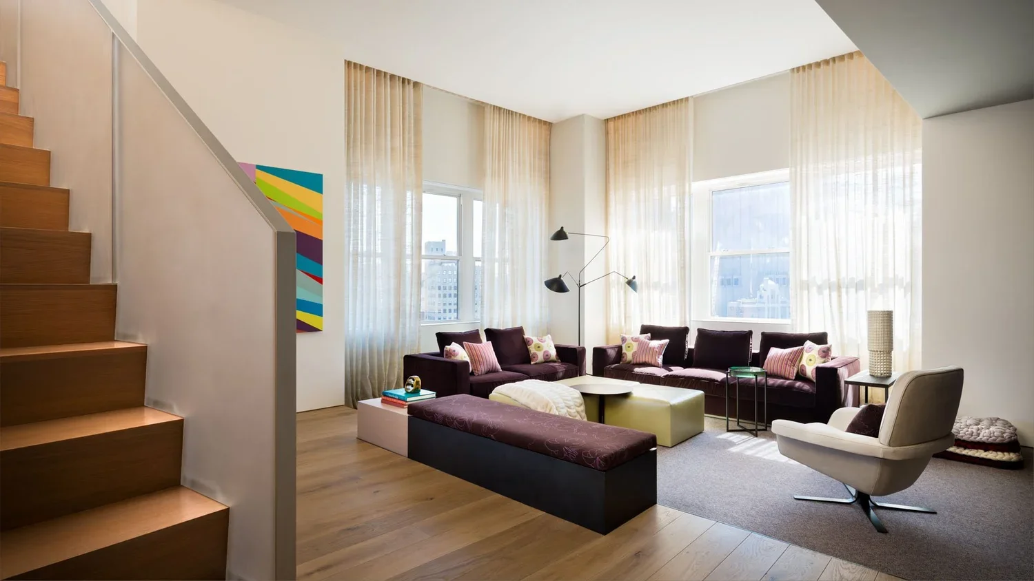 Bright living room with large windows, purple sofas with colorful pillows, a white armchair, a modern floor lamp, and light wood floors.