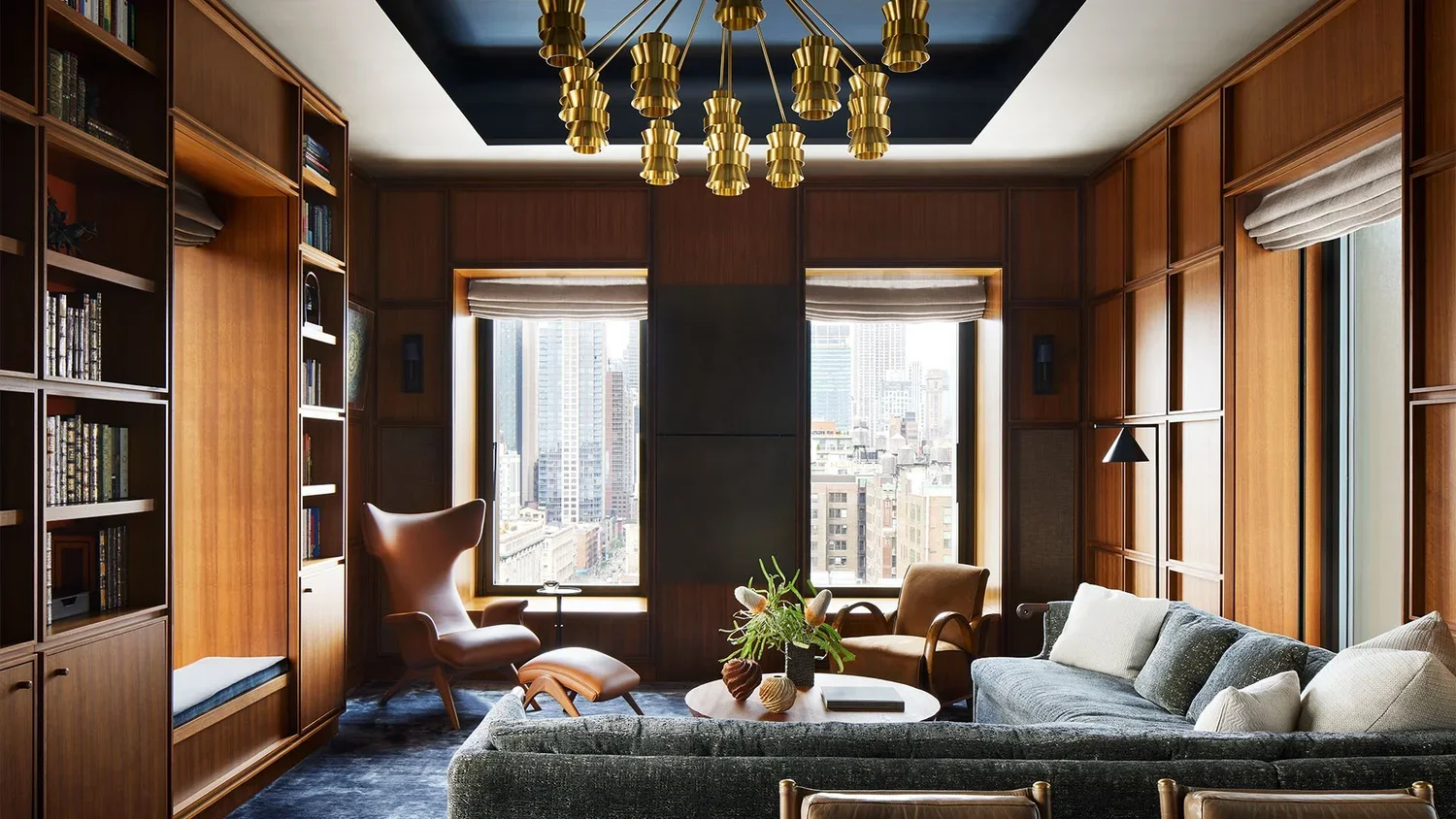 Modern living room with wooden wall panels, large windows with city view, a gray sofa, armchairs, a round coffee table with plants, books, and decorative objects, and a gold chandelier.