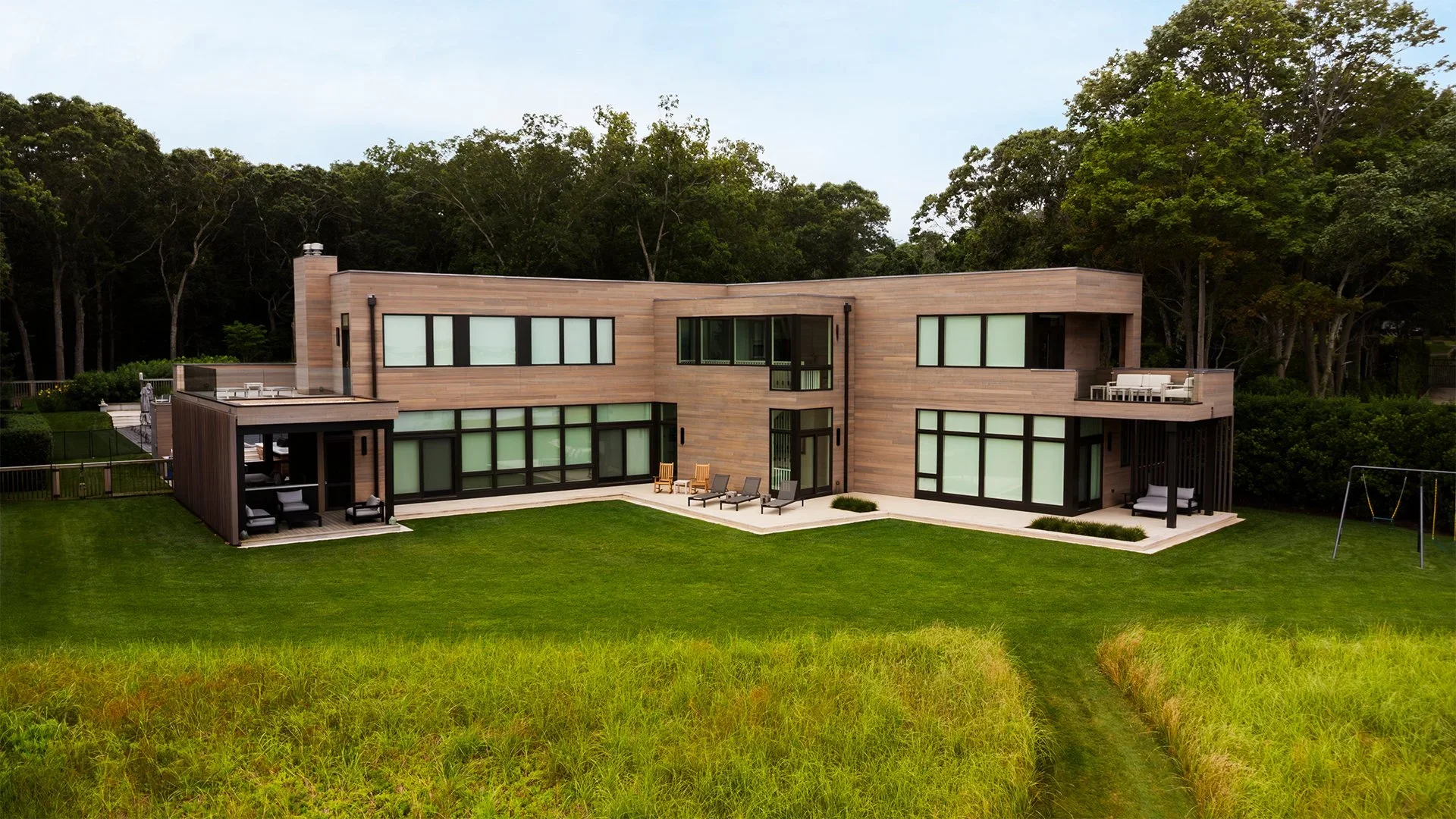 Modern two-story house with large windows, wooden exterior, green lawn, and surrounding trees.