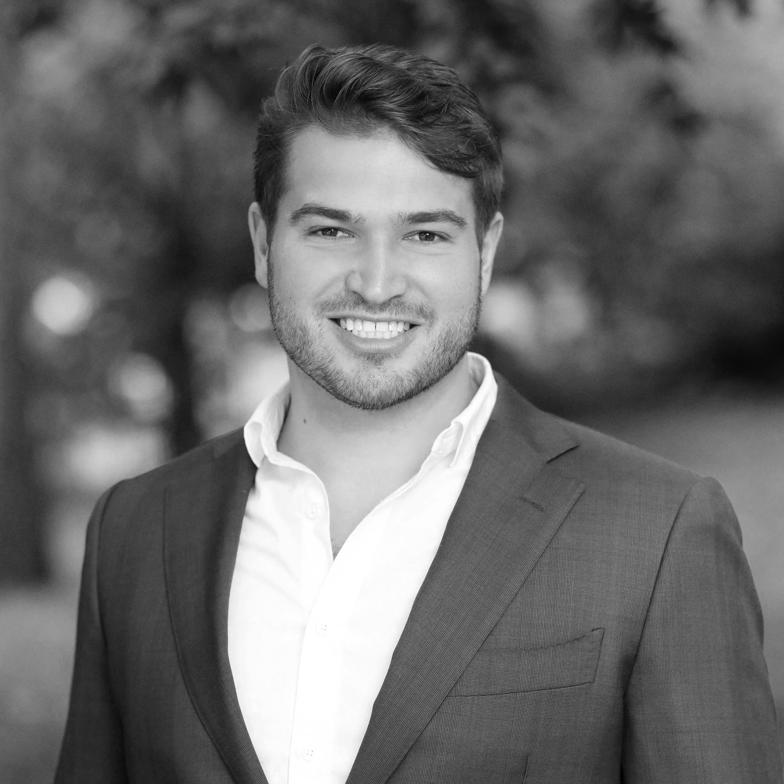 Black and white photo of a smiling charming young man wearing a suit against a tree lined background.