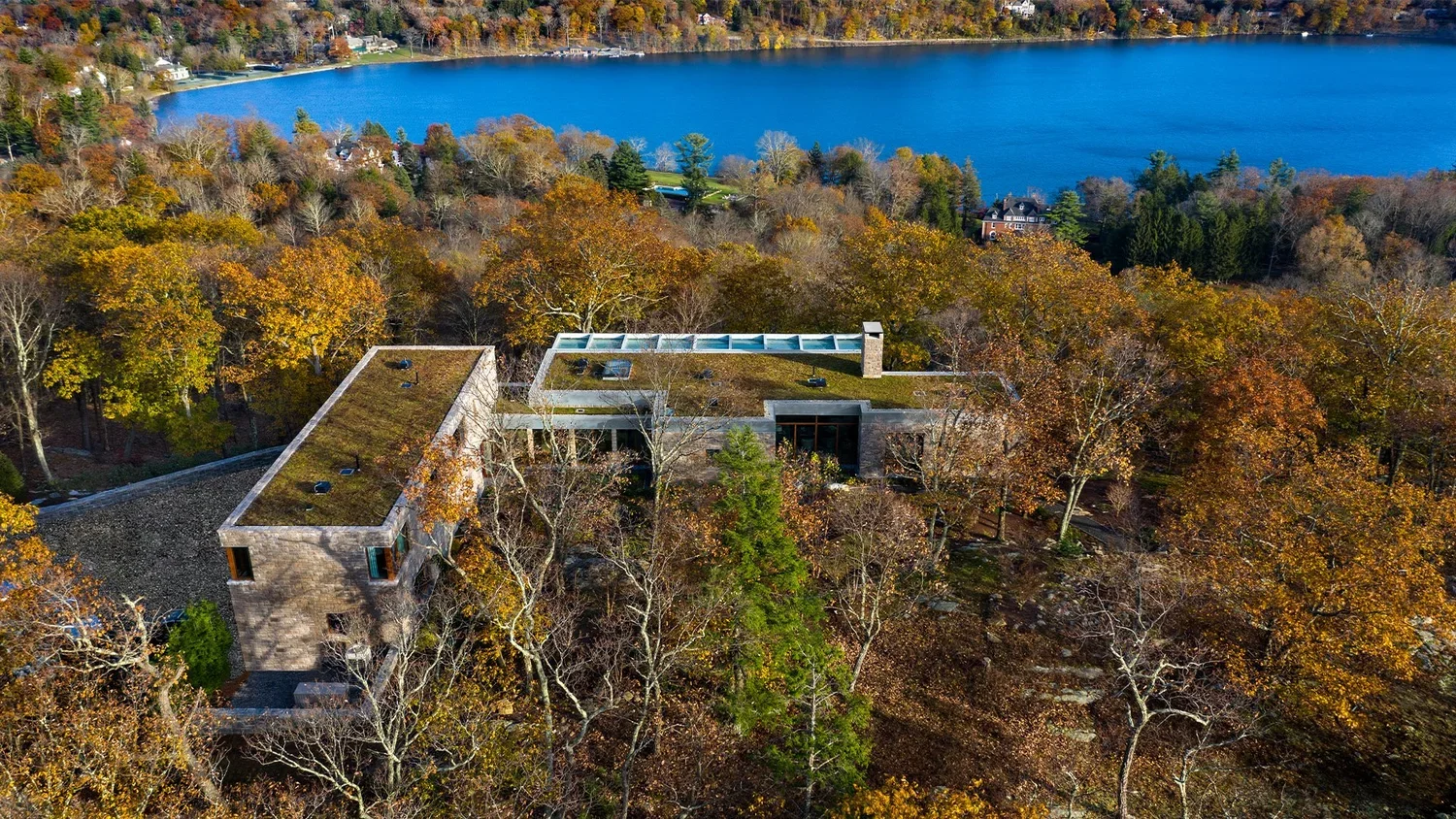 Aerial view of a modern house with a green roof amidst fall foliage, with a large blue lake in the background.