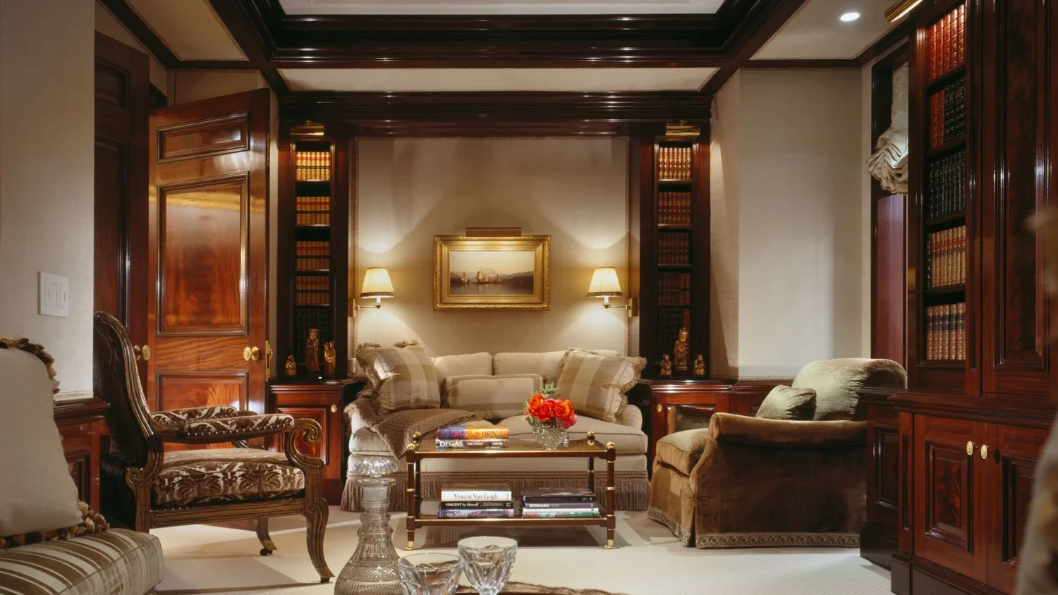 Elegant living room with cream-colored sofa, patterned armchair, and glass coffee table with books and a flower arrangement, surrounded by dark wood bookshelves and paneling, with a framed landscape painting and wall-mounted lamps.