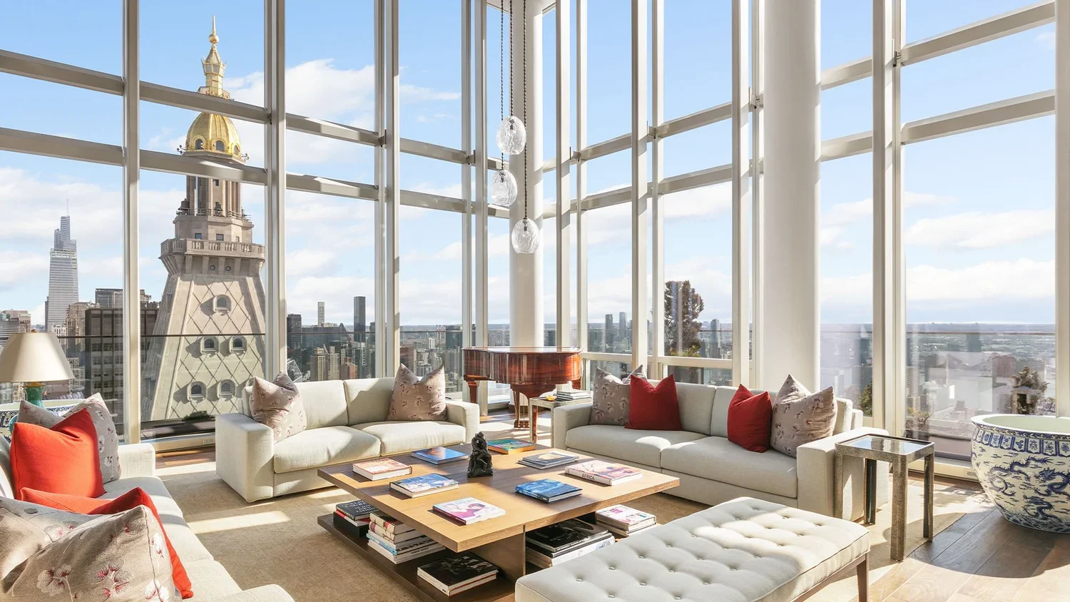 Luxury living room with large glass windows revealing a city skyline, including a distinctive tower with a gold dome, a grand piano, white sofas with red and floral pillows, a wooden coffee table with books, and a large decorative planter.