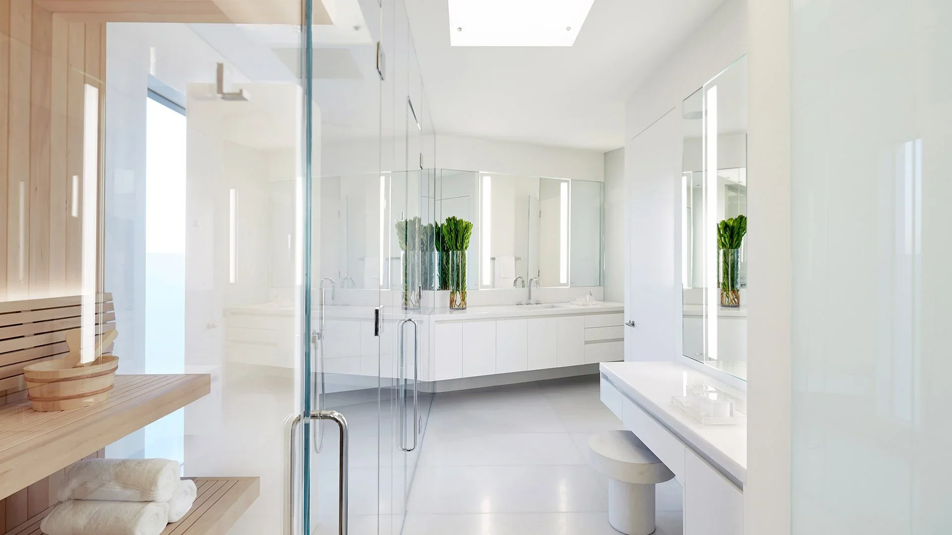 Modern bathroom with white walls, large mirror, and glass shower enclosure. Decorated with green plants in clear vases, featuring minimalistic white vanity and seating area.