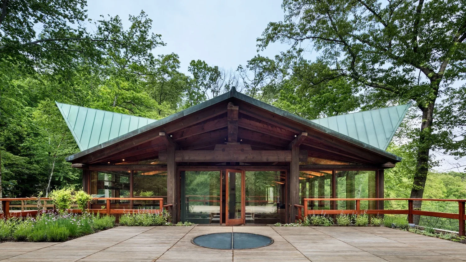 A modern glass house with a large deck, surrounded by trees and greenery, with a distinctive triangular roof.