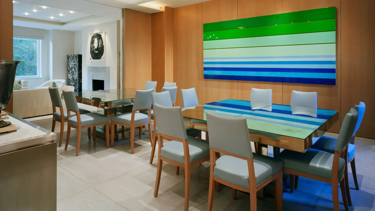 A modern dining room with a large glass top table surrounded by nine white chairs, a wooden wall with a colorful striped painting, and a cozy living room area with a white fireplace and artwork on the wall.