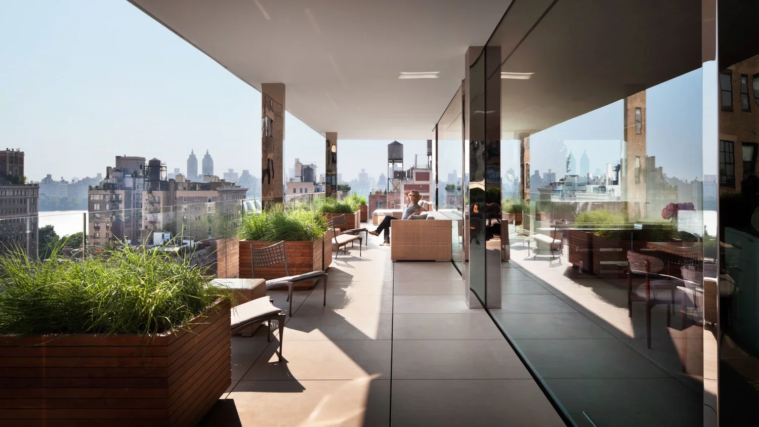 View of a modern rooftop terrace with seating, plants, and glass walls overlooking a city skyline with tall buildings.