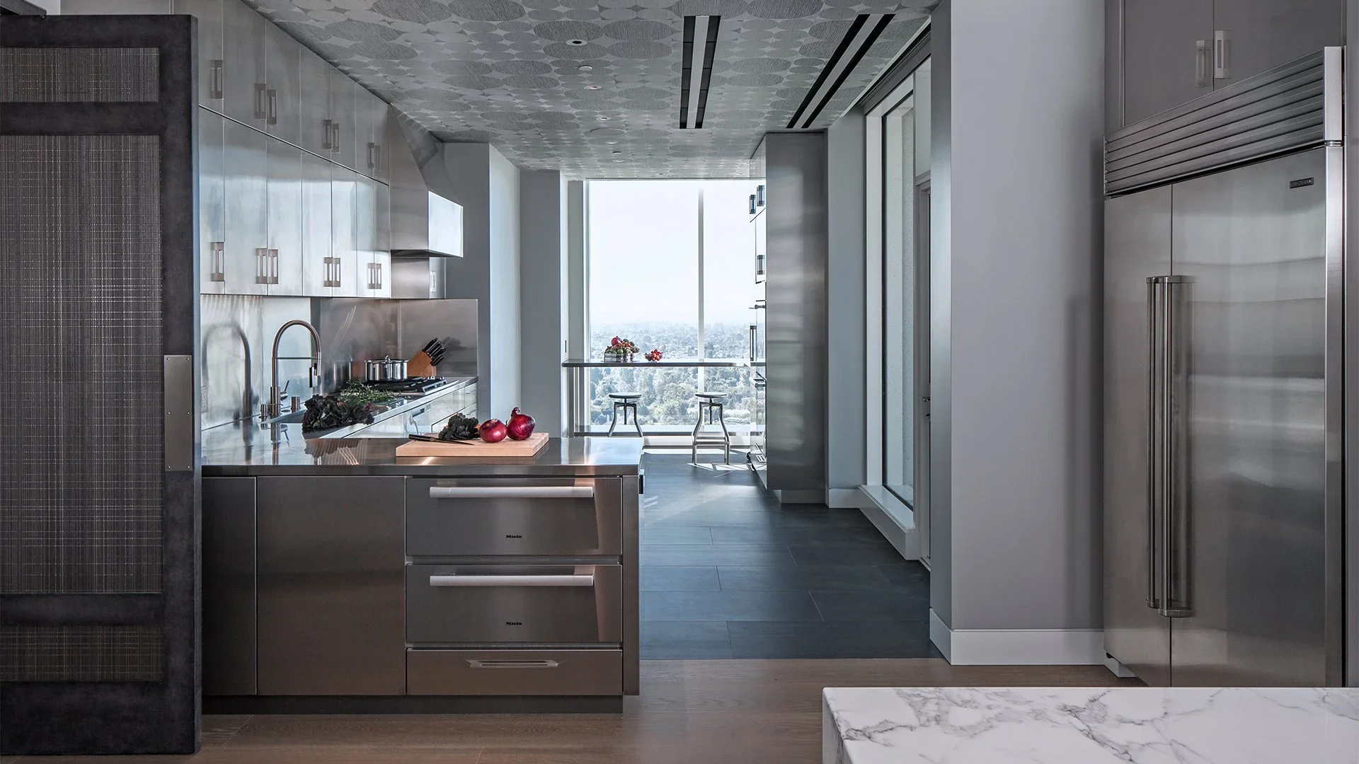 Modern kitchen with stainless steel appliances and large window overlooking cityscape, decorated with red onions and vegetables on the counter.