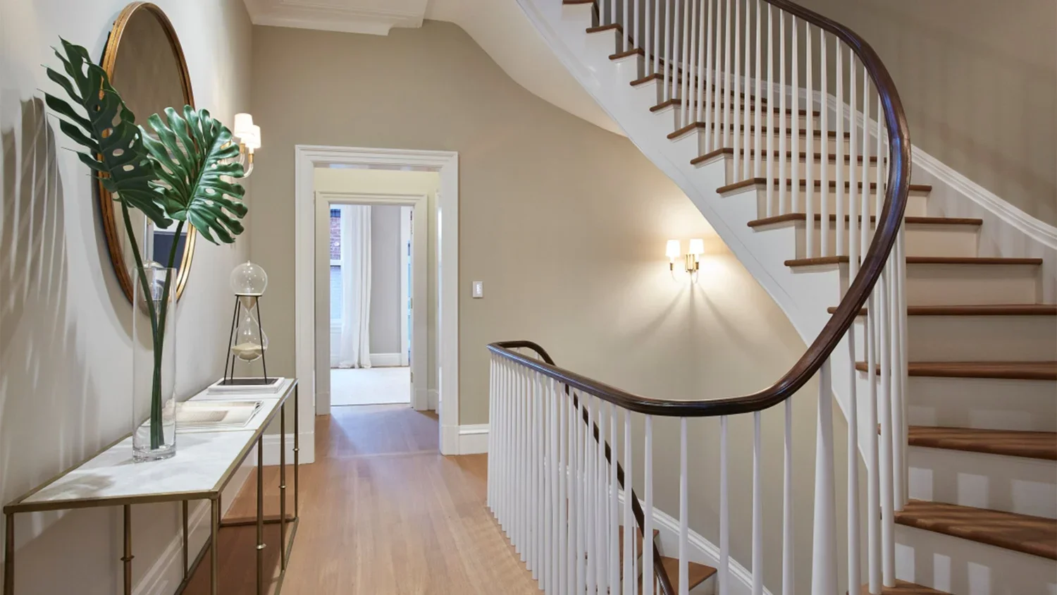 Interior view of a house entrance with a curved staircase, a console table with decorative items, and a large leafy plant as decor.