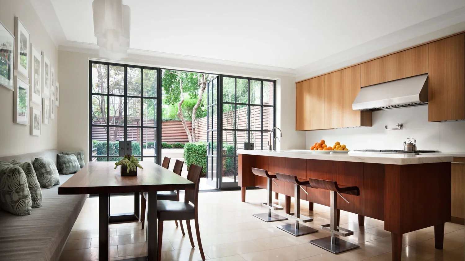 Modern kitchen and dining area with large glass doors opening to a backyard, featuring a wooden dining table with chairs and a kitchen island with bar stools, countertop with oranges, and a stainless steel range hood.
