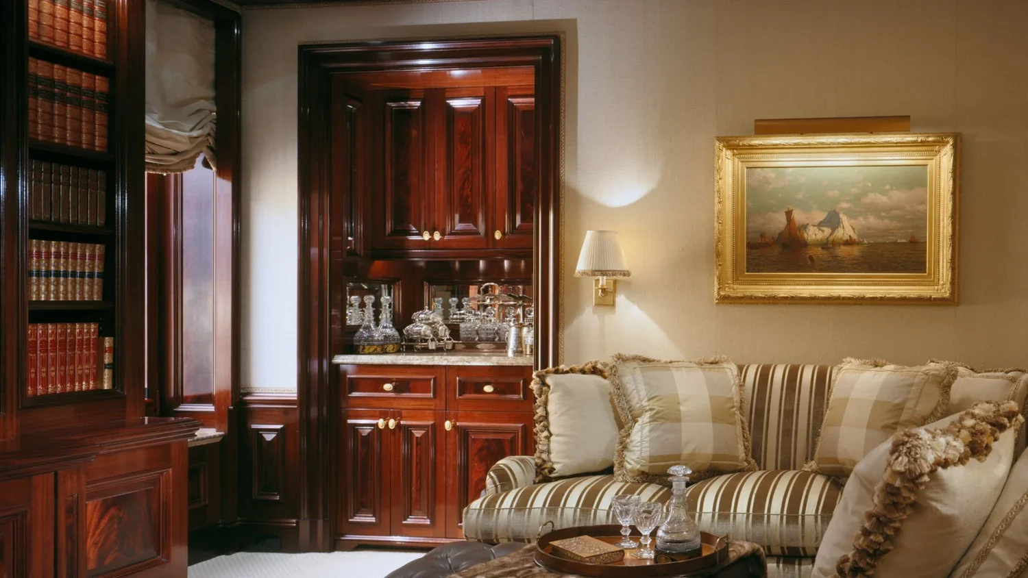 A cozy living room with a wooden cabinet, a striped sofa with decorative pillows, a painting of a ship on the wall, a wall sconce light, and a tray with glasses and a decanter on a table.