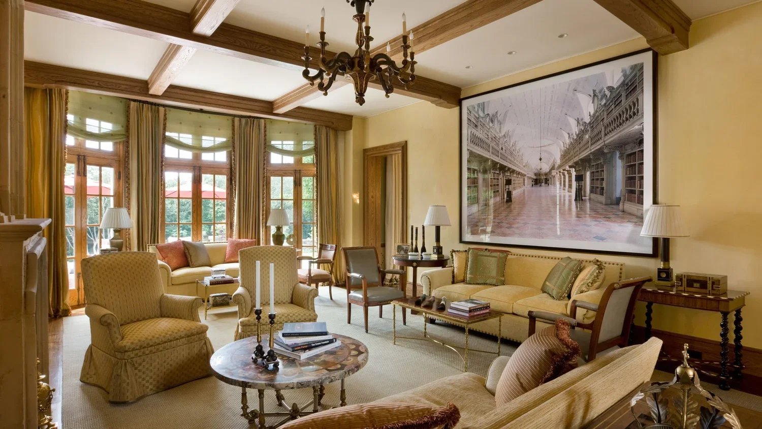Elegant living room with cream-colored sofa and armchairs, wooden beams on ceiling, large windows with yellow curtains, chandelier, and a large framed black-and-white photo of a library interior on the wall.