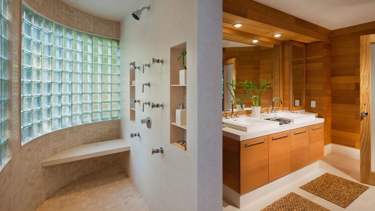 A bathroom with a corner shower area featuring glass block windows, multiple shower heads, and built-in shelves, separated from a double vanity with two sinks, a large mirror, and wooden cabinetry, all decorated with potted plants.