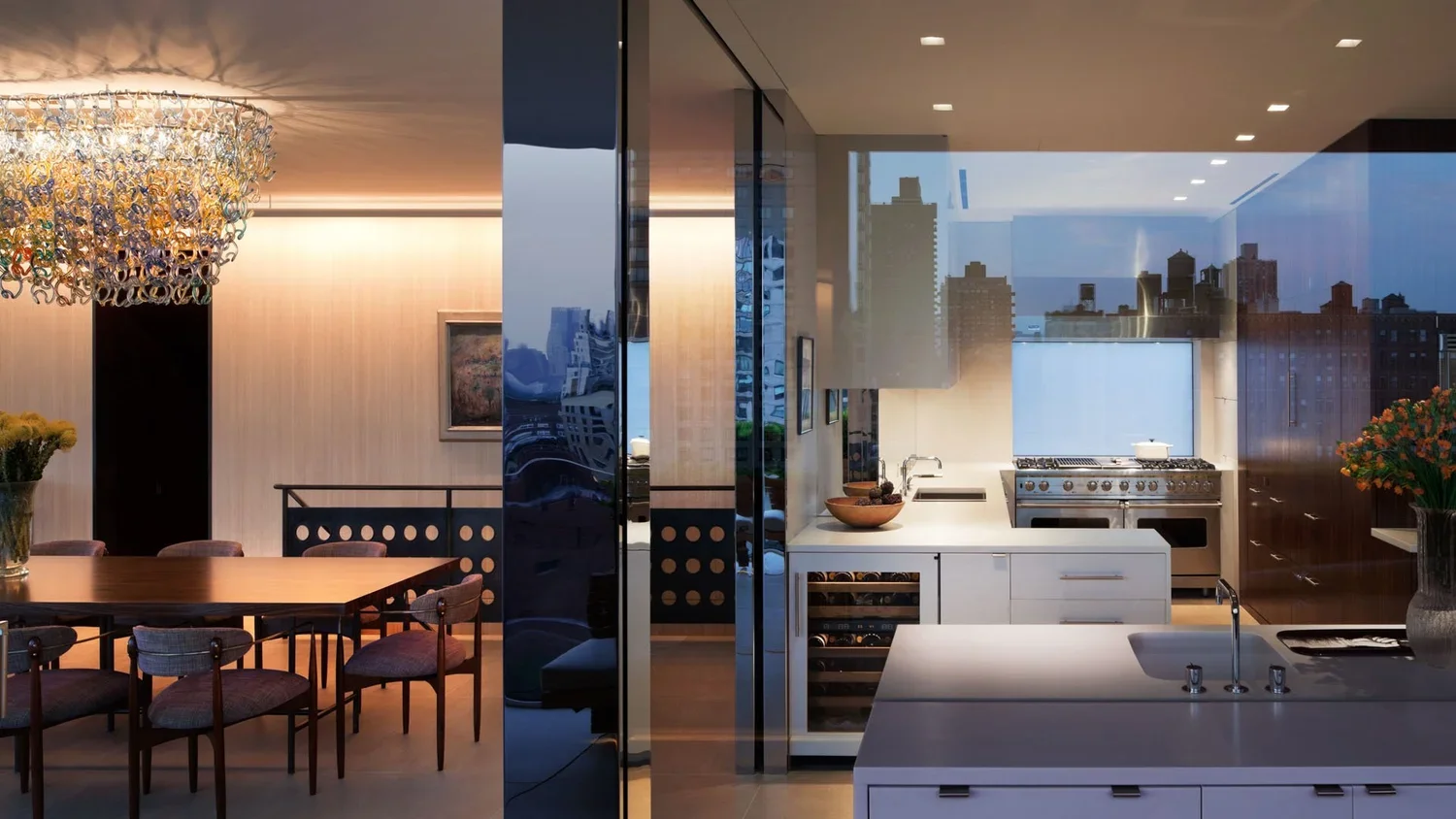 Modern kitchen and dining area with city skyline view through large windows, contemporary white cabinetry, stainless steel appliances, dining table with chairs, and decorative lighting.