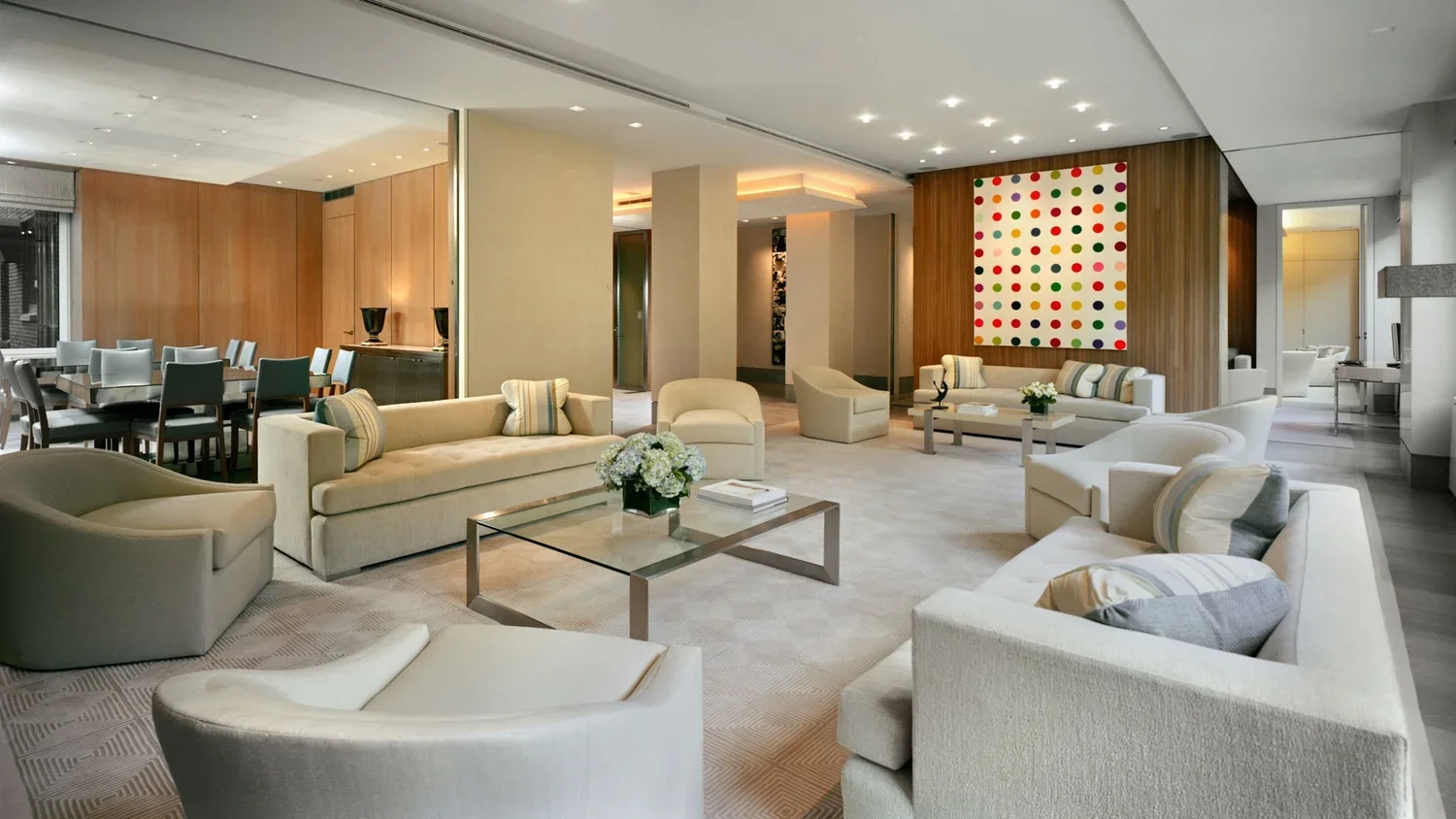Modern hotel lobby with cream-colored sofas, armchairs, a glass coffee table with a flower arrangement, a wooden-paneled wall with colorful abstract art, and a dining area in the background.