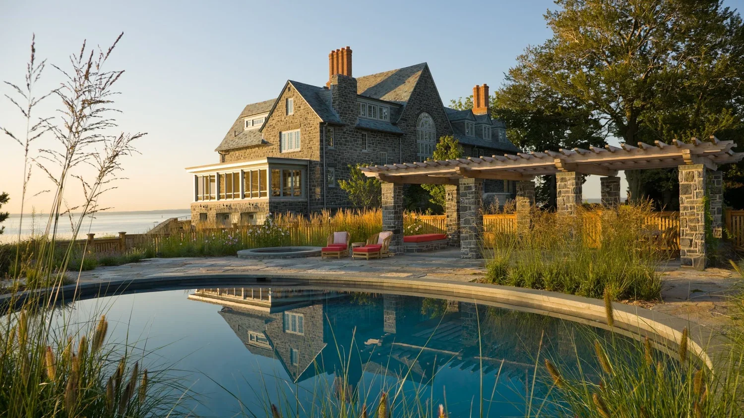 A large stone house with a pool in the foreground, a pergola with seating, and trees with a body of water in the background at sunset.
