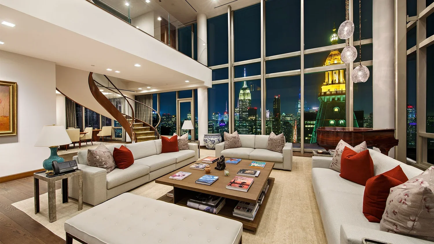 Living room with large windows showing a city skyline at night, featuring illuminated skyscrapers, with white sofas decorated with red and patterned throw pillows, a wooden coffee table with magazines, a grand piano, and a spiral staircase leading to