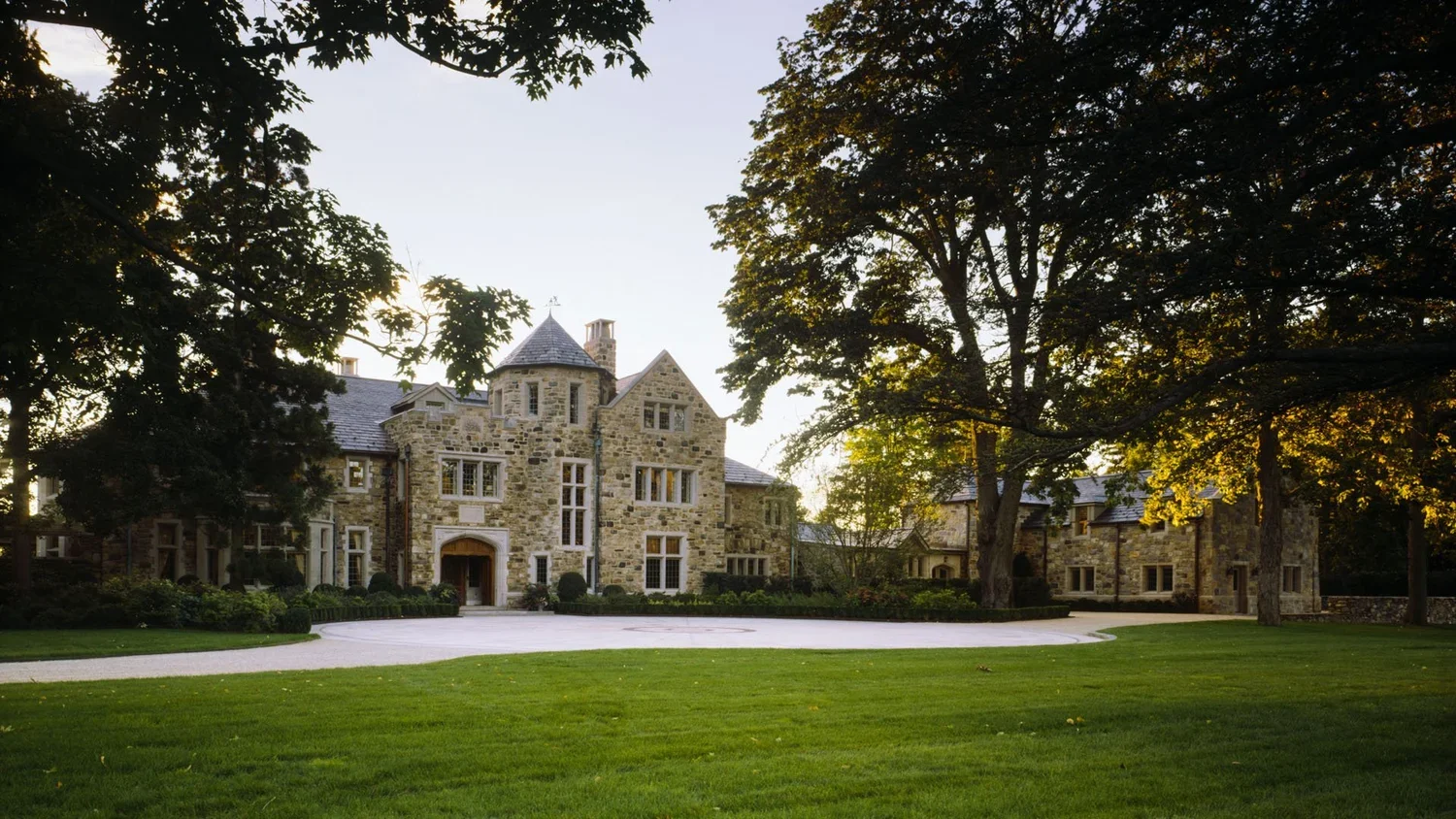 A large stone mansion with multiple gables, windows, and turrets, surrounded by tall trees and a well-maintained lawn in the front yard.