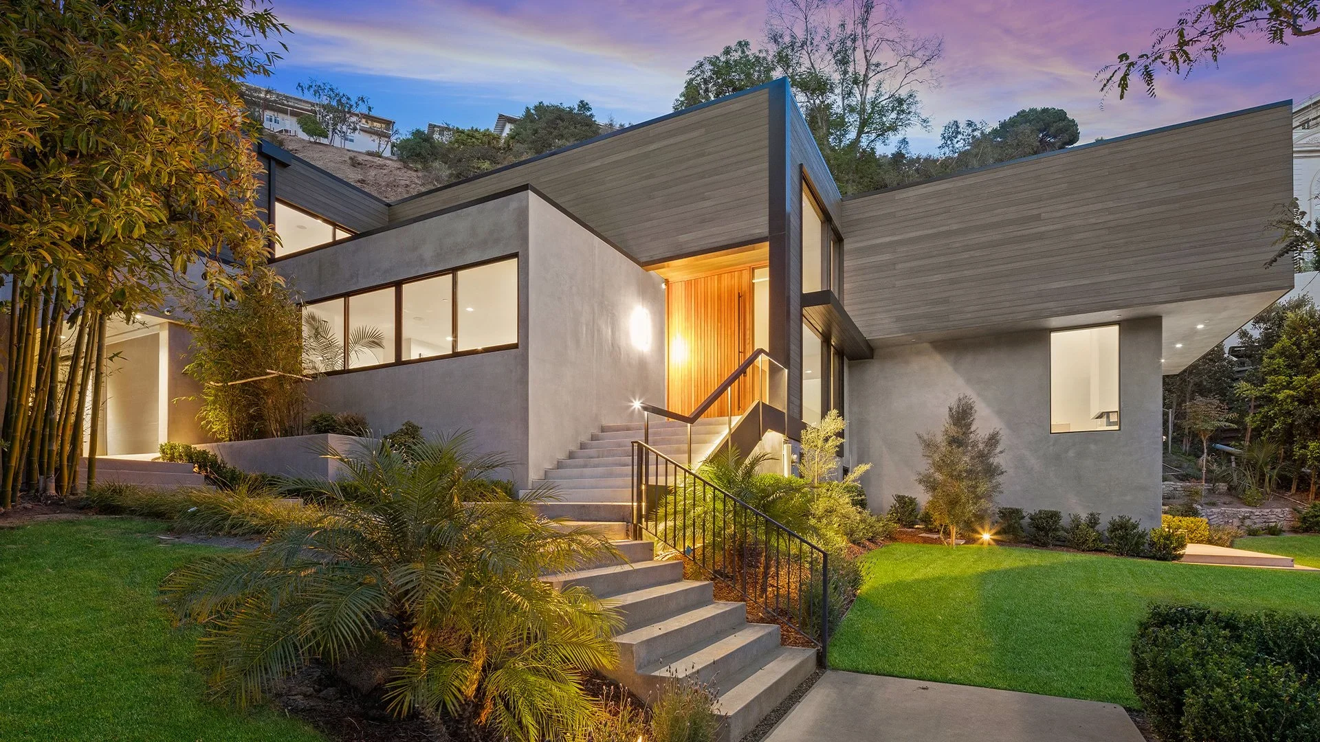 Modern house with gray concrete exterior, large windows, and landscaped front yard illuminated at dusk.