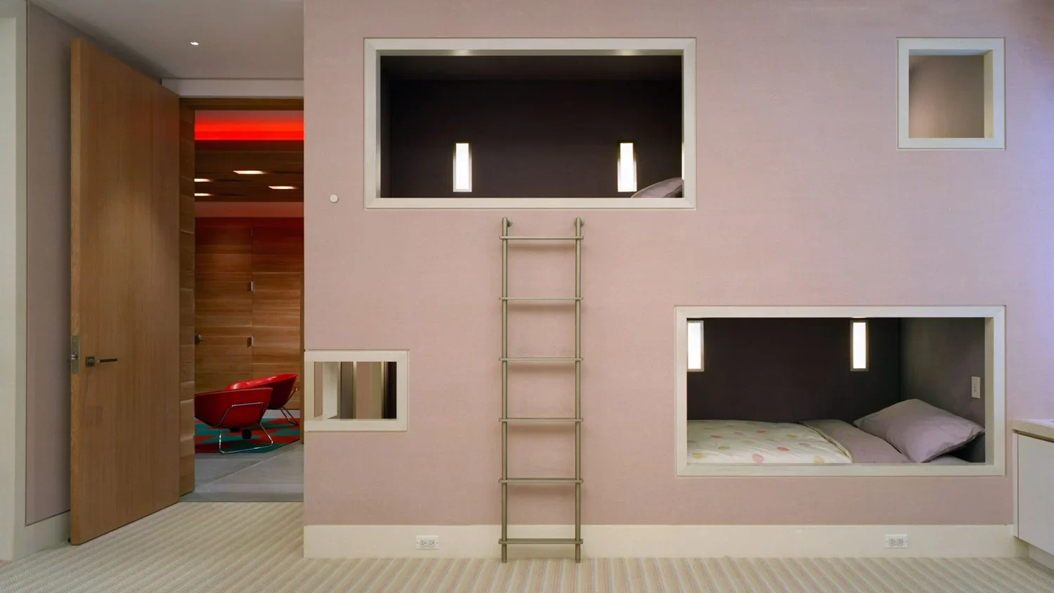 Room with two sleeping pods built into the wall, each with a small light and a pillow, a ladder leading to a loft above, and a doorway showing a red chair and wooden wall in another room.
