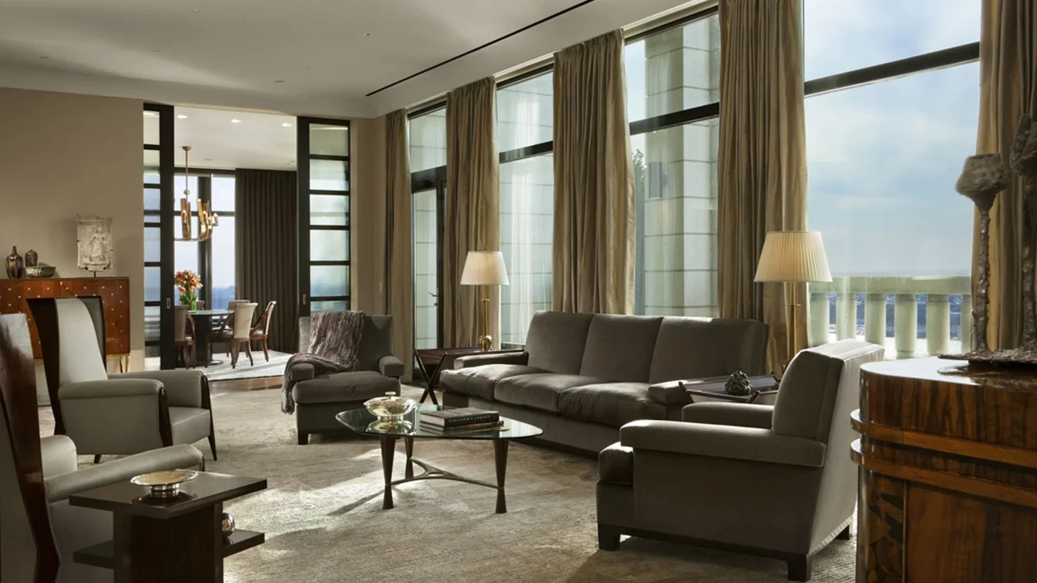 Spacious modern living room with large windows, beige curtains, gray sofas, armchairs, a glass coffee table, and table lamps, with a dining area visible in the background.