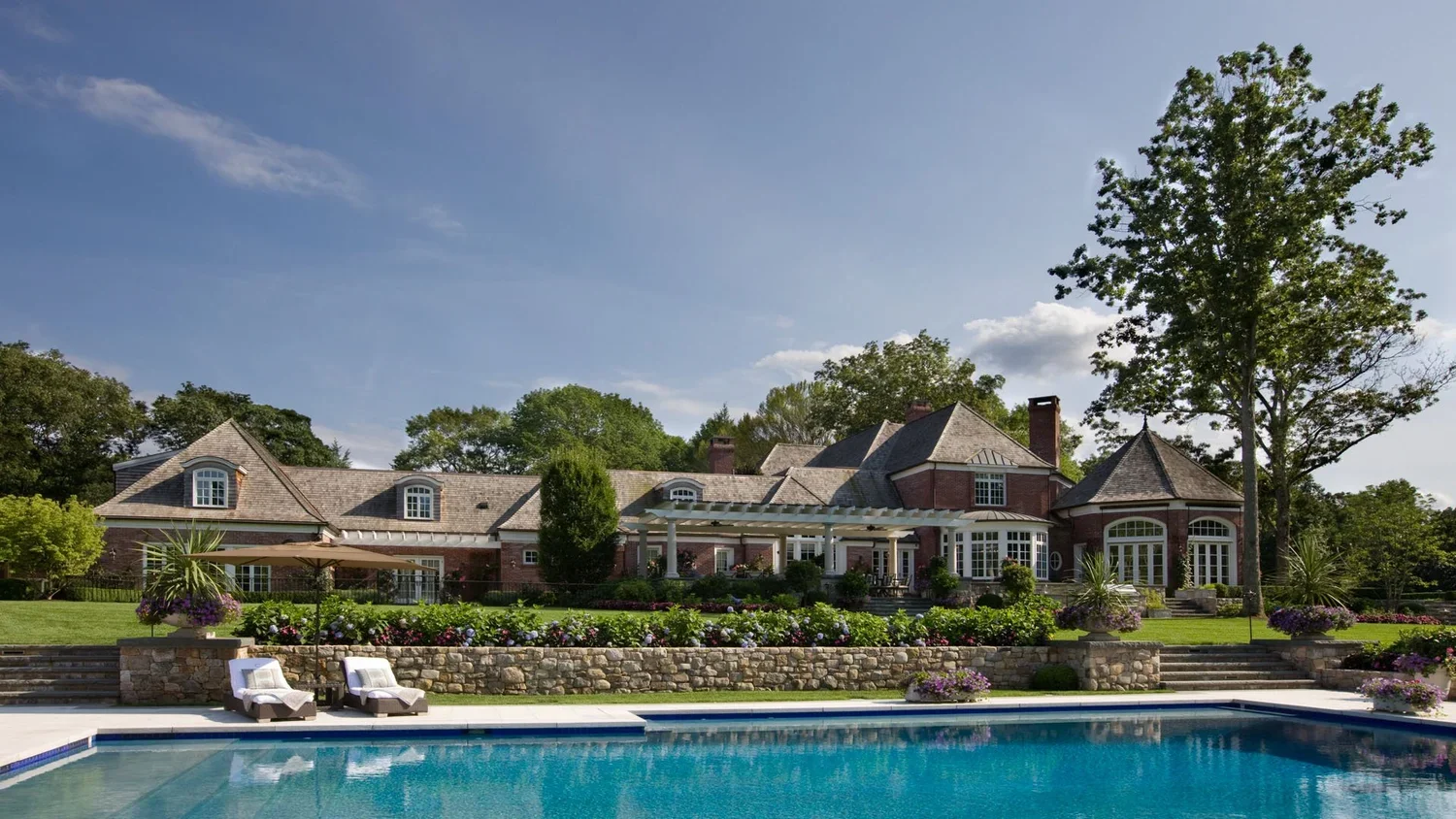 Luxury house with swimming pool in backyard, lush greenery, colorful flower beds, and a stone wall.