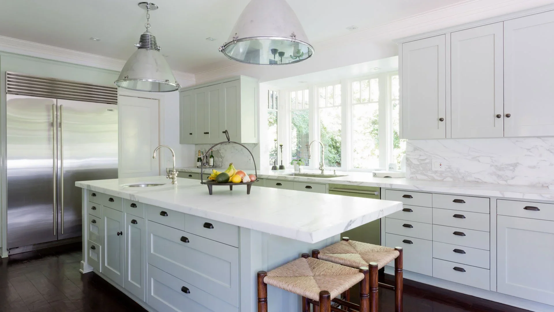 Bright kitchen with white cabinets, marble countertops, stainless steel refrigerator, large window, light fixtures, and two woven stools at the island.