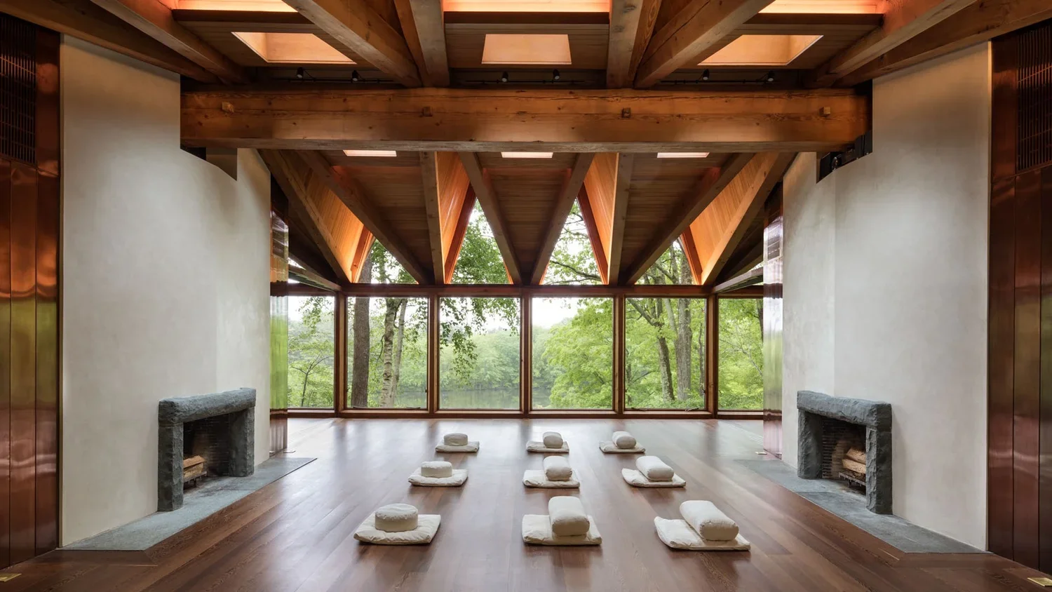 A spacious yoga studio with a wooden ceiling and large windows showing green trees outside. There are cushions and bolsters arranged on the floor for meditation or yoga practice, and two fireplaces on either side of the room.