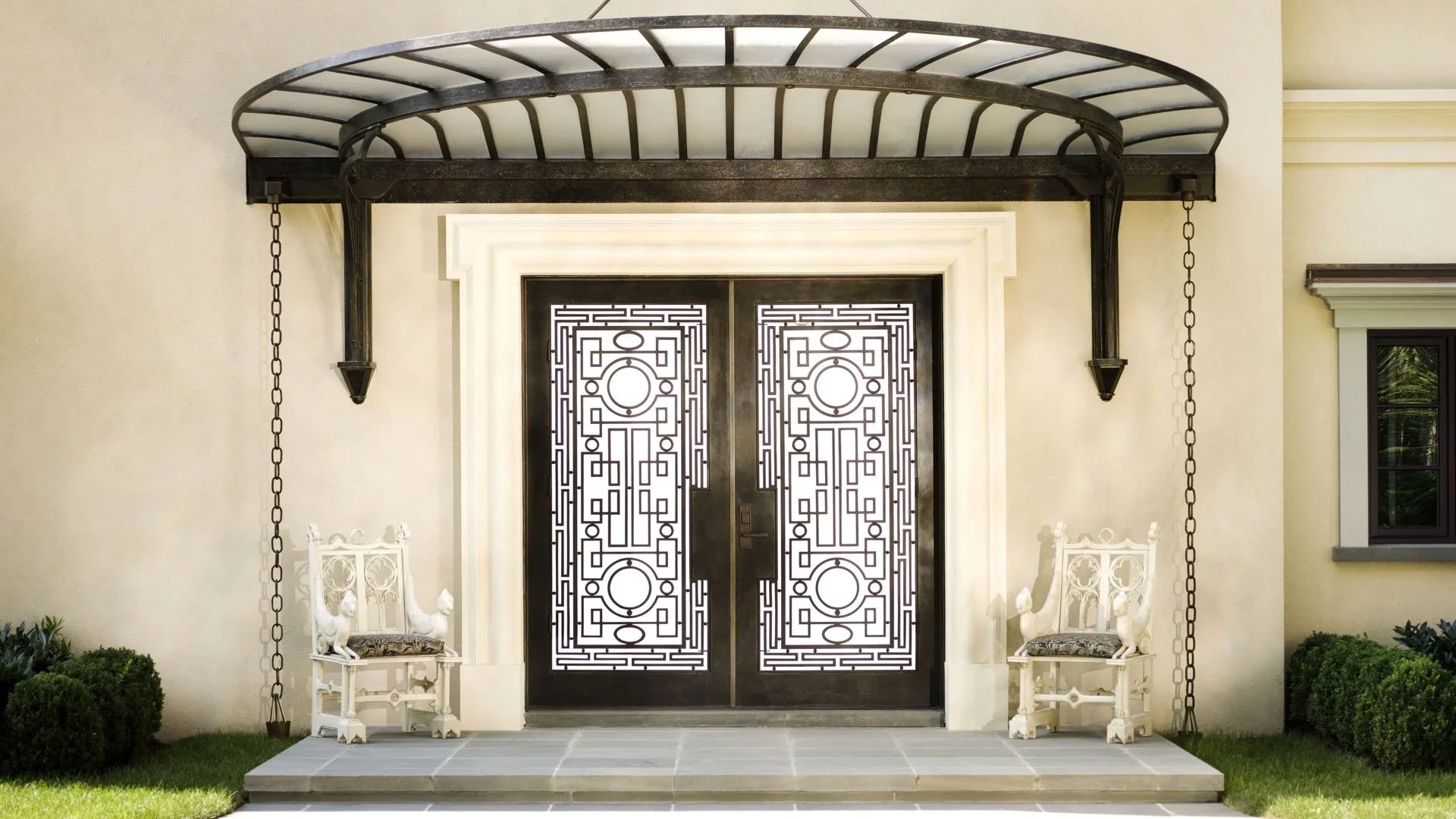 Black double doors with intricate geometric metalwork, framed by a white doorway, and flanked by two ornate white gates with lion statues on top, serving as armrests, and chain swings hanging from a decorative black canopy above the entrance. The are