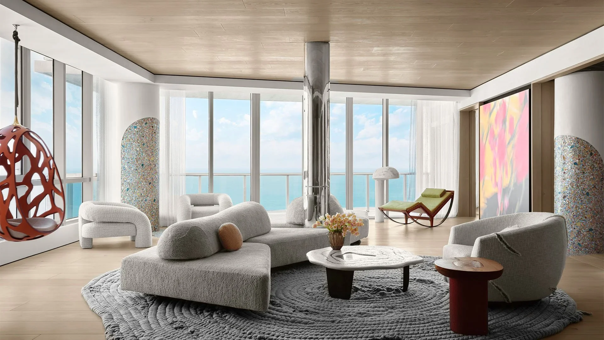 Modern living room with large windows offering a view of the ocean, featuring a gray sectional sofa, white armchairs, a round marble coffee table, a colorful textured headboard, abstract paintings, and various contemporary chairs and decor.
