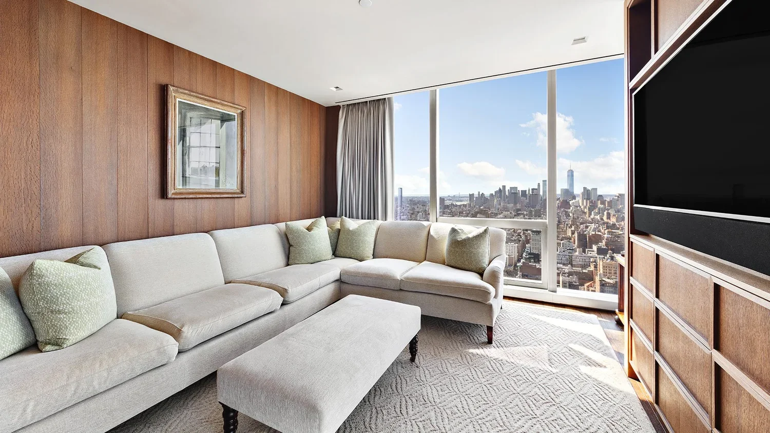 Living room with beige sectional sofa on a patterned rug, wood-paneled walls, large window with city skyline view, and a wall-mounted flat-screen TV.