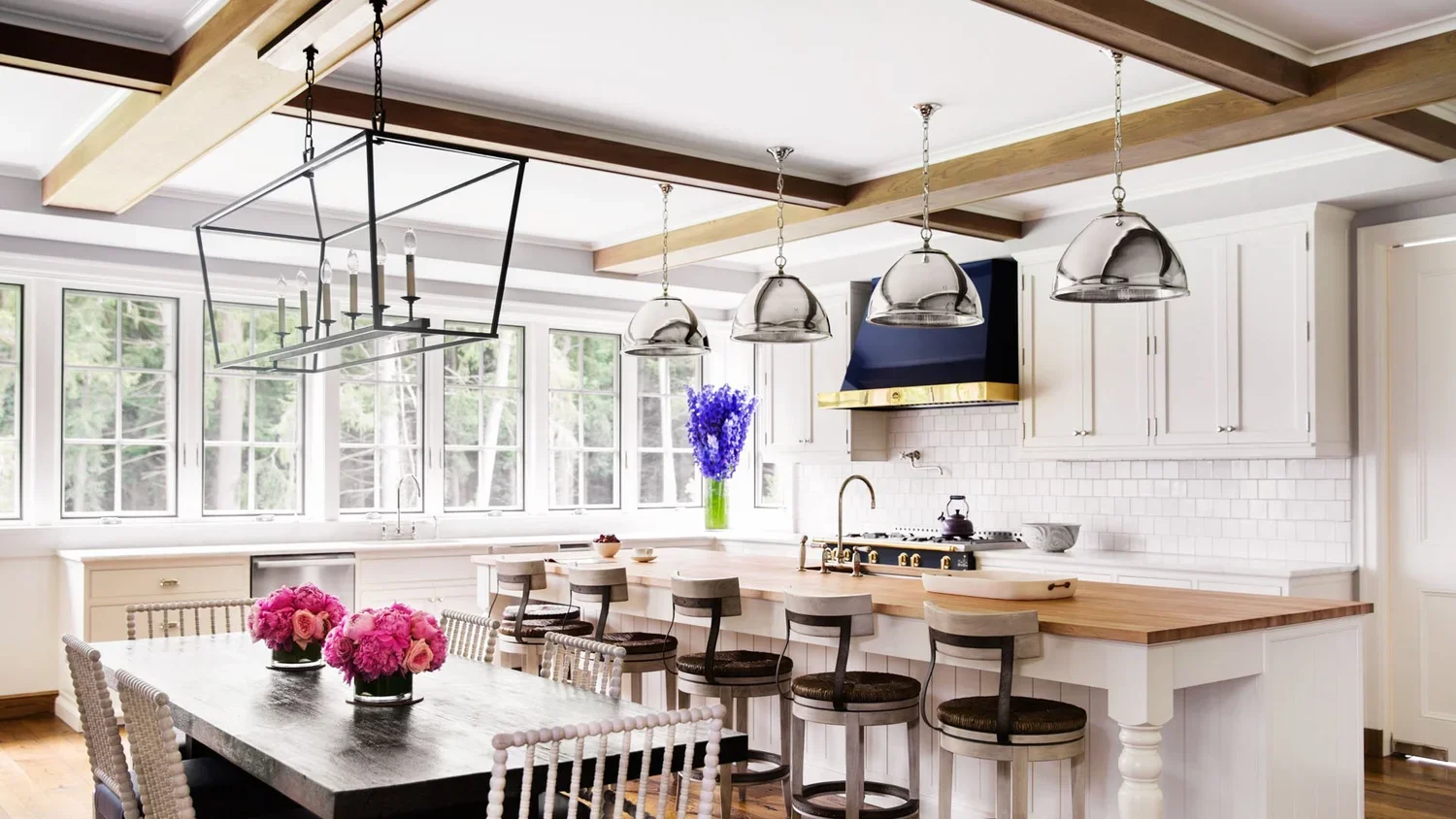 Bright kitchen with large windows, white cabinets, a wooden kitchen island with four chairs, a dining table with pink flowers in vases, and pendant lights hanging from beamed ceiling.
