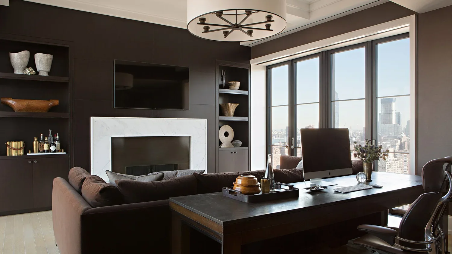 Modern living room with large windows showing city skyline, dark accent wall, fireplace with white marble surround, mounted flat-screen TV, bookshelf decor, brown sectional sofa, wooden coffee table, computer on desk, and decorative items.